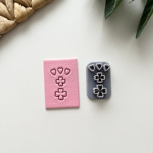 May include: A pink clay with heart and clover shapes pressed into it, next to a grey clay cutter with the same shapes. The shapes are for crafting and are arranged in a vertical pattern. The background is white.