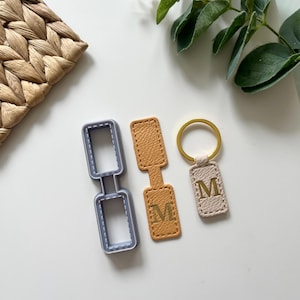 Op de afbeelding: Een verzameling leren sleutelhangers en een sleutelhanger-vormige koekjesuitsteker. De sleutelhangers zijn in de kleuren bruin, beige en goud, elk met de letter "M" in reliëf. De koekjesuitsteker is grijs.