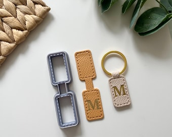 May include: A selection of leather keyrings and a keyring-shaped cookie cutter. The keyrings are in tan, beige, and gold colours, each with the letter "M" embossed. The cookie cutter is grey.