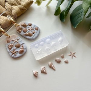 Può includere: Orecchini con sfondo azzurro, adornati con conchiglie e stelle marine nei toni del marrone e dell'oro. È presente anche uno stampo trasparente con forme di conchiglie e ciondoli singoli a forma di conchiglia, che suggeriscono un tema spiaggia o oceano.