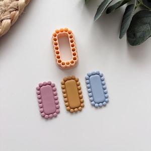 Pode incluir: Uma coleção de cortadores de biscoitos retangulares coloridos em tons de rosa, amarelo e azul, cada um com uma borda decorativa de contas. Um cortador de cor pêssego está posicionado acima dos outros. Os cortadores estão em uma superfície branca.