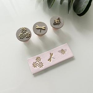 May include: Decorative stamps featuring floral, dragonfly, and bee motifs. The stamps have grey tops with gold-coloured embellishments and pink handles. A pink rectangular piece with matching designs is also included.
