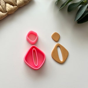 Puede incluir: Un juego de dos cortadores de galletas de plástico rosa en forma de rectángulo redondeado con un rectángulo más pequeño cortado en el centro. Los cortadores están sobre una superficie blanca junto a dos formas de arcilla marrón.
