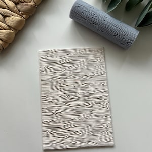 Puede incluir: Un rodillo de silicona blanco con un patrón de veta de madera. El rodillo está sobre una superficie blanca.