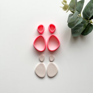 May include: Two pairs of polymer clay earrings. One pair is a light pink color and the other pair is a light gray color. The earrings are shaped like a teardrop with a rounded top and a pointed bottom.