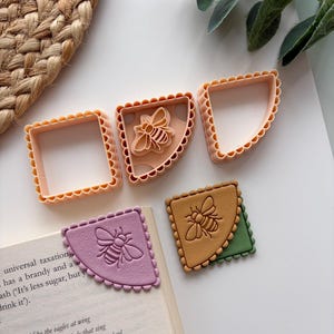 Op de afbeelding: Een verzameling roze koekjessnijders met geschulpte randen en twee hoekboekmarkeringen in de vorm van een bij. De bladwijzers zijn paars en bruin, met een groene achterkant. De koekjessnijders zijn vierkant en kwartcirkelvormig.