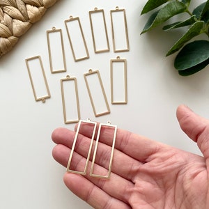 以下が含まれることがあります： 吊り下げ用の穴が上部に開いた、金色の金属製の長方形のフレームが10個セットになっています。フレームは白い表面に2列に並べて配置されています。