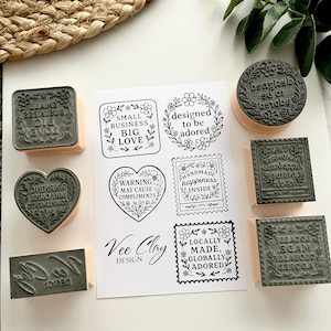 May include: A selection of rubber stamps with wooden mounts, featuring diverse designs. The stamps display floral patterns, hearts, and squares with phrases such as "Small Business Big Love" and "Locally Made, Globally Adored". A sheet of paper with corresponding designs is also visible.
