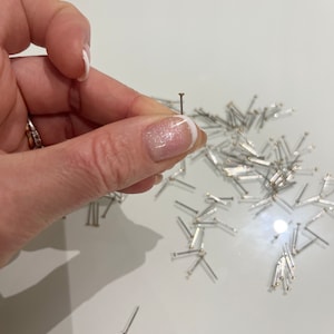 Op de afbeelding: Een hand houdt een enkele zilverkleurige speld vast met een kleine, ronde, goudkleurige kop. Veel vergelijkbare spelden liggen verspreid op een wit oppervlak. De spelden lijken nieuw en ongebruikt.