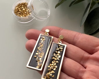 May include: A pair of black rectangular earrings with gold flakes embedded in the resin. The earrings have gold-colored hooks.