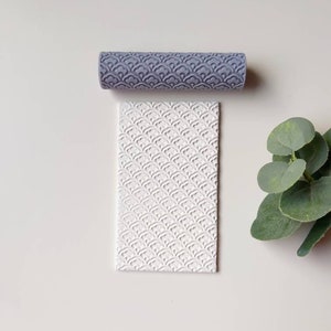 Pode incluir: Um rolo de massa cinzento com um padrão repetido de semicírculos sobrepostos. O rolo está ao lado de uma superfície branca com o mesmo padrão em relevo.