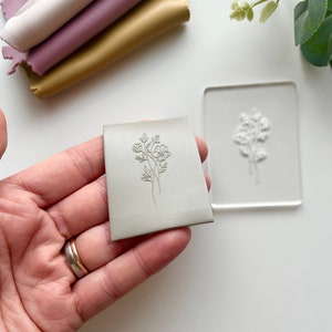 May include: A set of two gray polymer stamps with a floral design. The stamps are held in a hand wearing a silver ring.