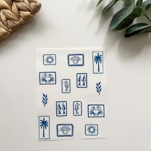 May include: A sheet of white stickers featuring blue illustrations of seaside motifs. The stickers depict a sun, seashells, palm trees, crabs, seahorses, and fish. The stickers are arranged in a grid pattern.