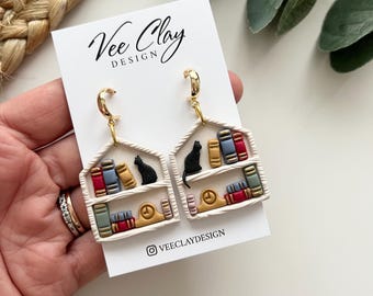 May include: A pair of gold hoop earrings with white bookshelf-shaped pendants. Each pendant features a black cat, colorful books, and a gold clock. The text "Vee Clay Design" and the Instagram handle are visible.
