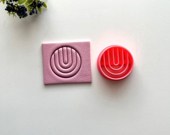Puede incluir: Una impresión de arcilla polimérica rosa de un círculo con un diseño en forma de U concéntrico y un cortador de galletas de plástico rojo con el mismo diseño.