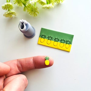 Könnte beinhalten: Nahaufnahme eines kleinen, gelben Ananas-förmigen Charms aus Polymer Clay mit grünen Blättern. Der Charm befindet sich auf einem Finger, und im Hintergrund ist eine Polymer Clay-Platte mit mehreren Ananas-Abdrücken zu sehen.