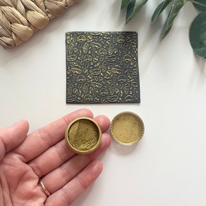 May include: Two small round containers of gold metallic paint, one open and one closed, on a white surface. A black sheet with a gold pattern of mushrooms and leaves is in the background.