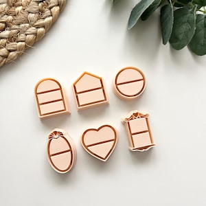 May include: Six light peach-coloured cookie cutters in different shapes, including a heart, oval, circle, and house. Each cutter has a simple design with a horizontal line or lines. The cutters are arranged on a white surface.