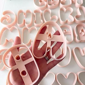 May include: A collection of light pink plastic cookie cutters shaped like letters of the alphabet. Each cutter features a raised edge for cutting and a central support structure. The letter A and the letter B are held in the foreground.