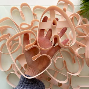 Może przedstawiać: Kolekcja jasnoróżowych plastikowych foremek do ciastek o różnych abstrakcyjnych kształtach. Foremki posiadają głęboką krawędź oraz centralną poprzeczkę zapewniającą wsparcie strukturalne. Kształty obejmują organiczne, płynne zarysy przypominające formy botaniczne.