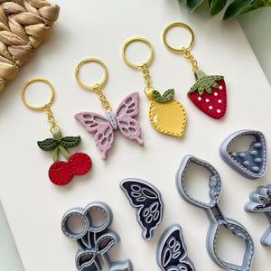 Op de afbeelding: Vijf leren sleutelhangers met verschillende bedels: een rode kers, een gele citroen, een roze vlinder, een rode aardbei en een grijze vlinder.  Ook afgebeeld zijn vijf grijze koekjesvormen in de vorm van een kers, een citroen, een vlinder, een aardbei en een vlinder.