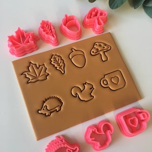 May include: A set of pink and brown plastic cookie cutters featuring autumnal themes, including leaves, acorns, mushrooms, a squirrel, a hedgehog, and a mug.