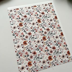 Könnte beinhalten: Eine rechteckige Karte mit floralem Muster. Das Design zeigt einen weißen Hintergrund mit einem sich wiederholenden Muster aus Blumen in Braun-, Rot- und Rosatönen sowie grünen Blättern und Stielen. Geeignet für Schreibwaren oder Grußkarten.