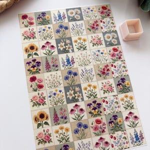 Può includere: Un foglio di adesivi con un design floreale patchwork, che mostra una griglia di fiori diversi. I fiori, tra cui girasoli e rose, sono in tonalità di rosa, giallo, viola e blu. È visibile anche un piccolo contenitore quadrato rosa chiaro.