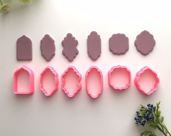 Puede incluir: Un conjunto de cortadores de galletas rosas en varias formas, incluyendo diseños arqueados, ovalados y festoneados. Arriba, se muestran recortes de color malva a juego. La disposición se establece sobre un fondo blanco, con acentos florales.