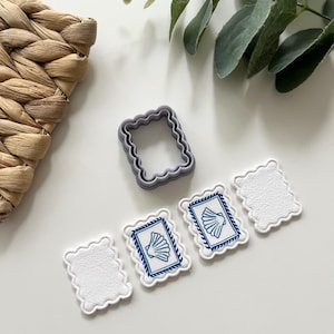 Könnte beinhalten: Ein grauer Ausstecher mit gewelltem Rand und drei weiße Dekorationen mit gewelltem Rand und blauen Muschelmotiven. Die Dekorationen sind auf einer weißen Oberfläche angeordnet, mit einem geflochtenen Korb und Grünzeug im Hintergrund.