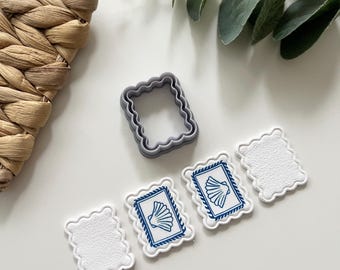 Pode incluir: Um cortador de biscoitos cinza com borda ondulada e três decorações brancas com borda ondulada e desenhos de conchas azuis. As decorações são dispostas sobre uma superfície branca, com uma cesta tecida e vegetação ao fundo.