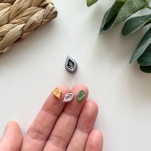 Könnte beinhalten: Drei kleine, blattförmige Stempel aus Polymer Clay in verschiedenen Farben: hellorange, helllila und hellgrün. Die Stempel befinden sich auf einer weißen Oberfläche.