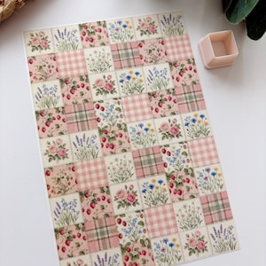 May include: A patterned paper sheet with a patchwork design. The design includes squares of pink gingham, plaid, and floral patterns. The floral patterns feature roses, strawberries, and wildflowers. A small, light pink square container is also present.