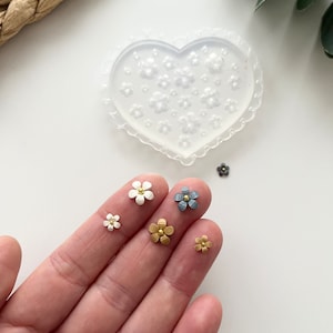 Puede incluir: Cinco pequeños amuletos de flores de resina en diferentes colores. Los amuletos son blancos, dorados y grises. Los amuletos están en una mano, y un molde de silicona en forma de corazón está en el fondo.