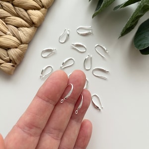 Op de afbeelding: Een close-up van een hand die zilveren oorhaakjes vasthoudt. De haakjes zijn klein en hebben een gladde, afgeronde vorm. Er liggen nog een aantal andere haakjes verspreid op een wit oppervlak.