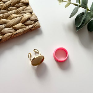 Op de afbeelding: Een goudkleurige verstelbare ring met een ovale zetting en een apart roze, cirkelvormig onderdeel. De ring ligt op een wit oppervlak, met een geweven strooien mat en groen in de achtergrond. De ring is ontworpen om een cabochon te bevatten.