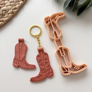 Puede incluir: Un conjunto de tres artículos: dos llaveros de cuero color terracota con forma de bota vaquera y un cortador de galletas a juego. Los llaveros tienen costuras detalladas y recortes florales. El cortador de galletas es de color melocotón.