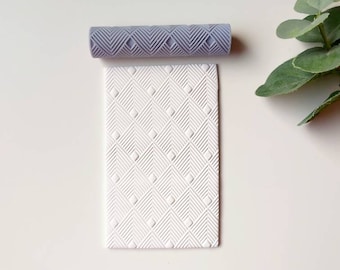 May include: A gray and white patterned rolling pin and a white embossed fondant sheet. The rolling pin has a geometric pattern of diamonds and lines. The fondant sheet has a similar pattern embossed into it.