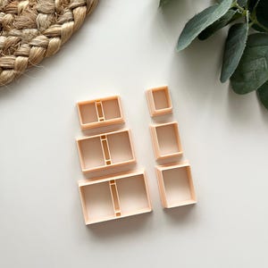 May include: Six light peach-coloured square cookie cutters in varying sizes. The cutters are plastic and have a rectangular shape, some with a central divider. They are arranged on a white surface, suitable for baking.
