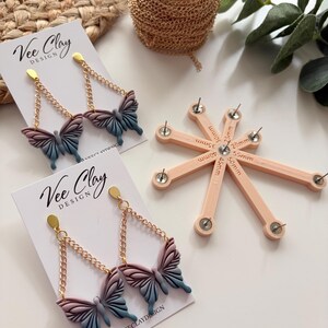 May include: Butterfly-shaped earrings with gold-colored chains and posts. The butterfly wings are a gradient of pink, purple, and blue. The earrings are displayed on white cards with the text "Vee Clay Design". A spool of gold chain and a pink tool are also visible.