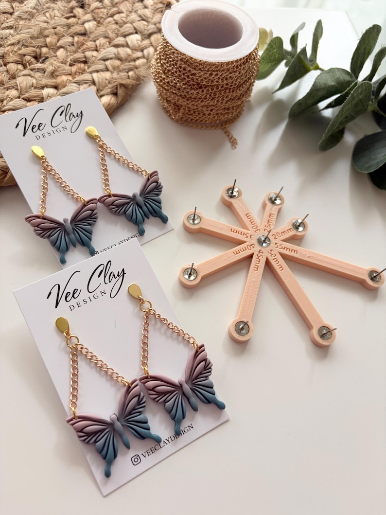 May include: Butterfly-shaped earrings with gold-colored chains and posts. The butterfly wings are a gradient of pink, purple, and blue. The earrings are displayed on white cards with the text "Vee Clay Design". A spool of gold chain and a pink tool are also visible.
