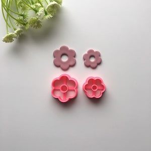 May include: Two pink plastic flower-shaped cookie cutters and two pink plastic flower-shaped fondant cutters. The cutters are on a white surface.