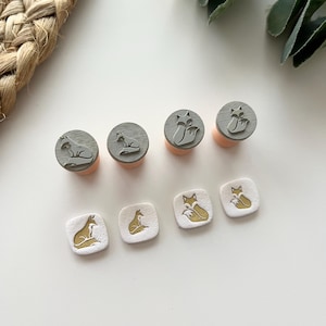 May include: A selection of wooden stamps and square embellishments with fox designs. The stamps have grey tops and pink bases. The embellishments are white with gold fox illustrations. The fox designs vary in pose.