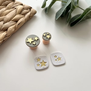 Könnte beinhalten: Eine Sammlung handgefertigter Ohr-Gauges und -Stecker mit Stern-Design. Die Gauges sind grau und goldfarben, während die Stecker rosa und goldfarben sind. Das Stern-Design ist auf der Oberfläche geprägt. Die Stecker sind rund, und die Gauges sind quadratisch.