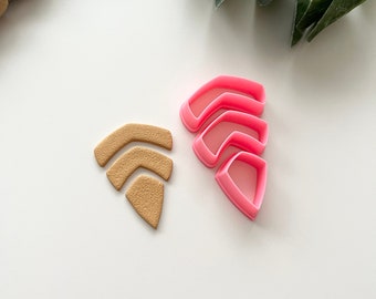 Può includere: Un set di cinque tagliabiscotti in plastica rosa a forma di una serie di archi sovrapposti. I tagliabiscotti sono mostrati accanto a un biscotto tagliato a forma di archi, di colore marrone e con una texture.