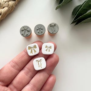 May include: A selection of bow-shaped stamps. Three grey, round stamps with bow designs are shown alongside three white, square stamps with gold bow designs. The stamps are on a white surface.