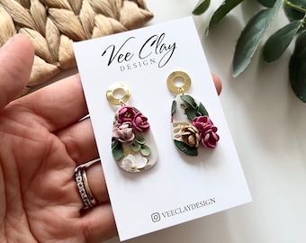 May include: A pair of white teardrop-shaped earrings with pink and gold floral accents. The earrings are on a white card with the text "Vee Clay Design" and the Instagram handle "@veeclaydesign".