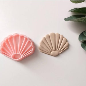 Puede incluir: Un cortador de galletas de plástico rosa en forma de concha y un molde de galletas de concha beige. Ambos están detallados con crestas y una forma redondeada.