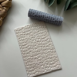 May include: A white textured sheet of faux leather with a crocodile pattern. A smaller, gray textured sheet of faux leather with a crocodile pattern is rolled up next to it.