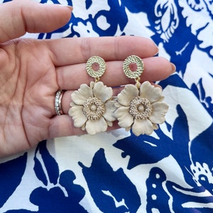 May include: A pair of cream-colored flower dangle earrings with gold-toned metal accents. The earrings feature a delicate flower design with a circular center.